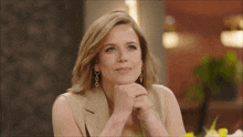 a woman wearing a tan jacket and gold earrings is sitting with her hands folded