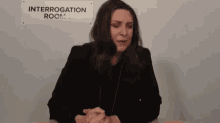 a woman in a black jacket is sitting at a table in front of a sign that says interrogation room .