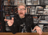 a man with glasses and a beard is sitting at a table in front of a shelf full of dvds