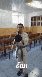 a young boy in a karate uniform is holding a black object in front of chairs and a sign that says ban on it