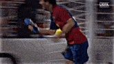 a man in a red shirt and blue shorts is running in a boxing ring with the uefa logo in the background .