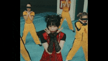 a woman in a red dress and black gloves stands in front of a group of women in yellow pants
