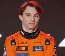 a young man with red hair is wearing an orange racing suit with logos for android and cisco