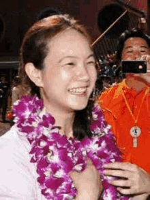 a woman wearing a lei of purple flowers is smiling while a man takes a picture .