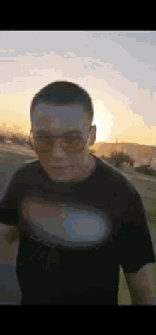 a man wearing sunglasses and a black t-shirt is walking down a street at sunset .