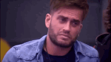 a man with a beard is wearing a blue denim jacket and a black shirt and making a sad face .