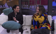 two women are sitting on a couch talking to each other . one of the women is wearing a colorful sweater .