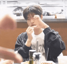 a young man sitting at a table with his hands on his face and a foreign language written on the bottom right corner
