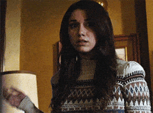 a woman wearing a sweater with a pattern on it stands in front of a lamp