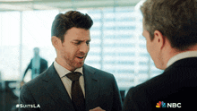 a man in a suit and tie is talking to another man in a suit and tie with the nbc logo behind him