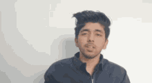 a young man in a blue shirt is standing in front of a white wall and making a funny face .
