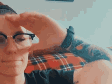 a woman wearing glasses and a watch is sitting on a plaid couch covering her face with her hand .