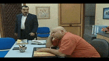 a bald man sits at a desk with his head resting on his hand while a man in a suit stands behind him