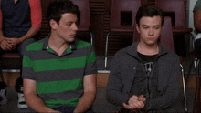 a man in a green and gray striped shirt sits next to another man in a gray jacket