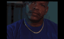 a bald man wearing a blue shirt and gold chains looks at the camera
