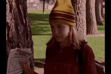 a young girl wearing a yellow and red beanie is standing next to a tree holding a flyer .
