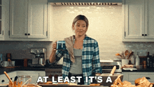 a woman in a plaid shirt is holding a cup of coffee in a kitchen with the words at least it 's a