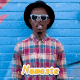 a man wearing sunglasses and a plaid shirt is standing in front of a blue brick wall and says namaste