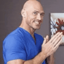 a bald man wearing a blue scrub top is clapping his hands in front of a painting .