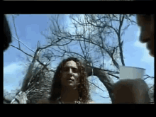 a man with long curly hair stands in front of a tree