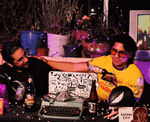 two men sitting at a table with a sign that says satan city on it