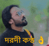 a man with a beard wearing sunglasses and a black shirt with a yellow thumbs up sign below him in a foreign language