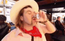 a man wearing a cowboy hat and a red bandana drinks from a glass