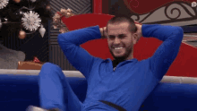 a man in a blue jumpsuit sits on a couch with his hands behind his head .