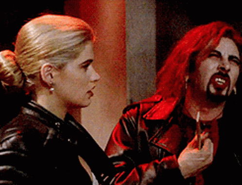 A blonde woman in a black leather jacket stares at a campy vampire feigning 