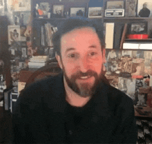 a man with a beard is smiling while sitting in front of a bookshelf filled with pictures .