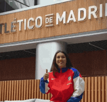 a woman giving a thumbs up in front of a sign that says " atletico de madrid "