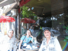 two men and a woman sit at a table in front of a coca cola umbrella