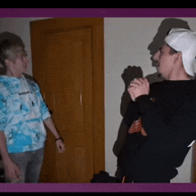 two young men are standing next to each other in a room . one of the men is wearing a tie dye sweatshirt .