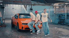 three men are dancing in front of an orange car with a sign that says wax shampoo full detail