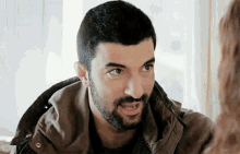 a man with a beard is wearing a brown jacket and talking to a woman