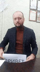 a man in a suit sits at a table holding a sign that says привет