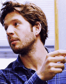 a man with a beard wearing a blue plaid shirt looks to his left