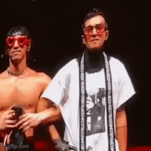 two shirtless men are standing next to each other on a stage . one of the men is wearing sunglasses and a scarf .
