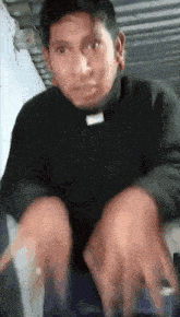 a man wearing a black sweater and a priest collar is sitting down with his hands on his knees .