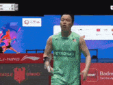 a man wearing a green petronas shirt stands in front of a badminton advertisement