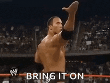 the rock is standing in a wrestling ring with his fist in the air and the words `` bring it on '' behind him .
