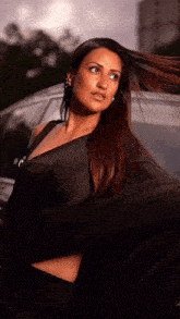 a woman in a black dress is standing in front of a car with her hair blowing in the wind