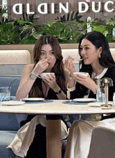 two women are sitting at a table in front of a sign that says " plain duc "