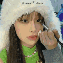 a woman wearing a fur hat and a green shirt is applying makeup to her face .