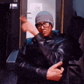 a man wearing glasses and a beanie is sitting in a chair and making a funny face .