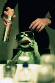 a kermit the frog is brushing his teeth in a bathroom while wearing sunglasses .