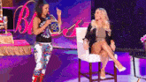 a woman is standing next to another woman who is sitting in a chair on a stage .