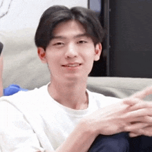 a young man in a white shirt is sitting on a couch with his hands folded and smiling .