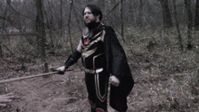 a man in a black cape is holding a stick in a forest