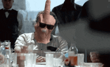 a man sitting at a table with glasses and a hand giving the middle finger